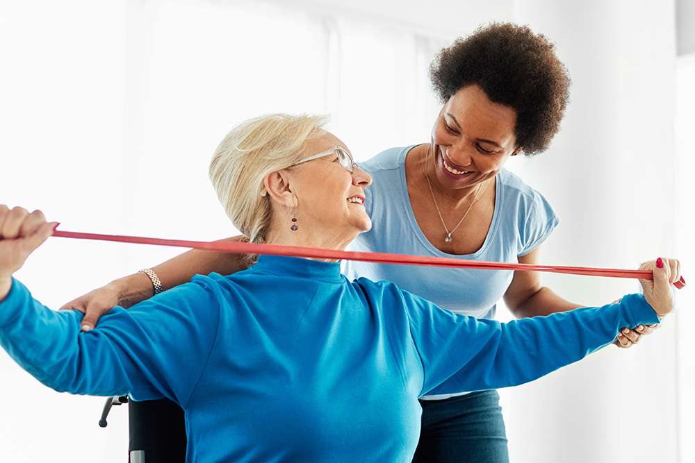 Senior woman in a senior living community doing physical therapy with a caregiver using a resistance band.