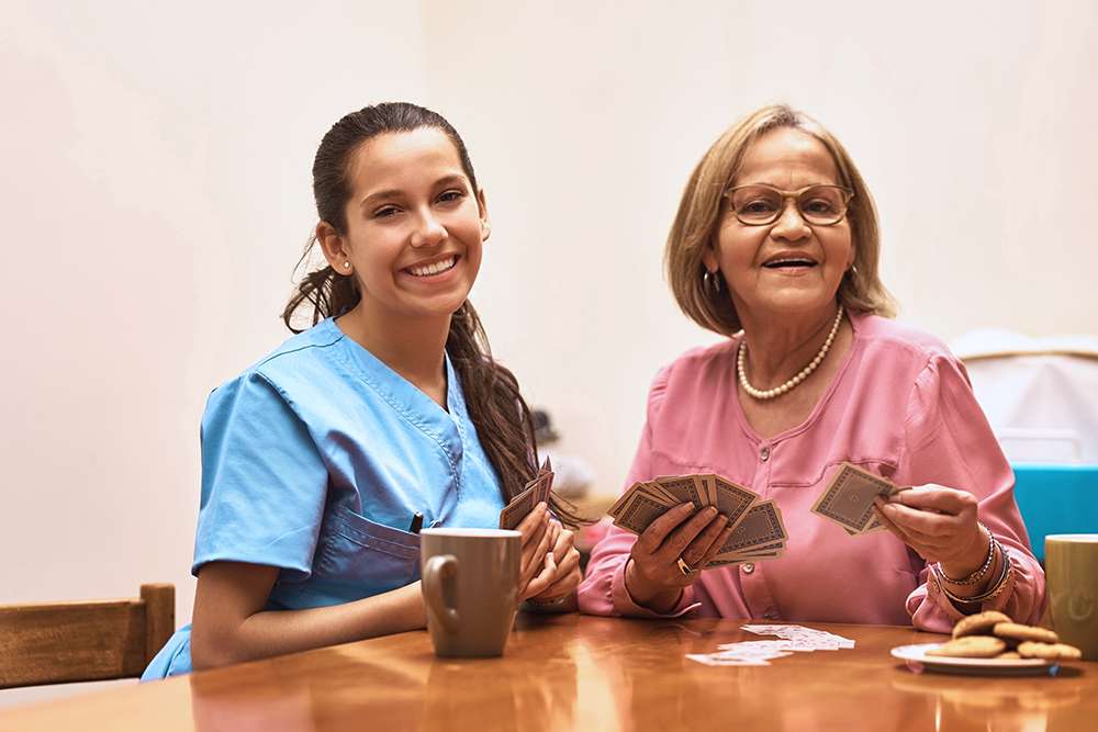 Caregiver and senior woman playing cards at a table in a senior living community.